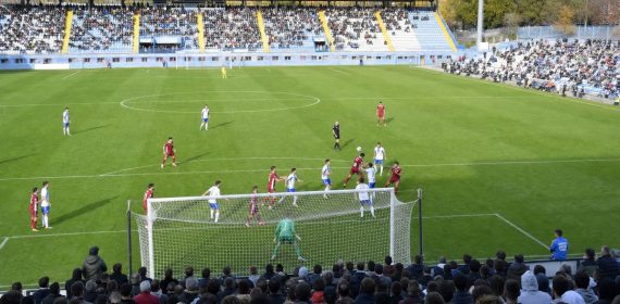 Dos futbolistas saltan a por un balón dividido en el área durante un partido en un estadio con la grada a medio llenar
