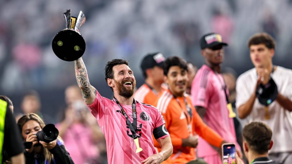 Un futbolista con camiseta rosa, brazalete de capitán y medalla levanta un trofeo negro y plateado mientras sonríe
