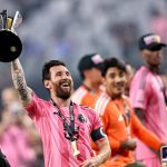 Un futbolista con camiseta rosa, brazalete de capitán y medalla levanta un trofeo negro y plateado mientras sonríe