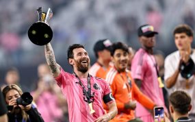 Un futbolista con camiseta rosa, brazalete de capitán y medalla levanta un trofeo negro y plateado mientras sonríe