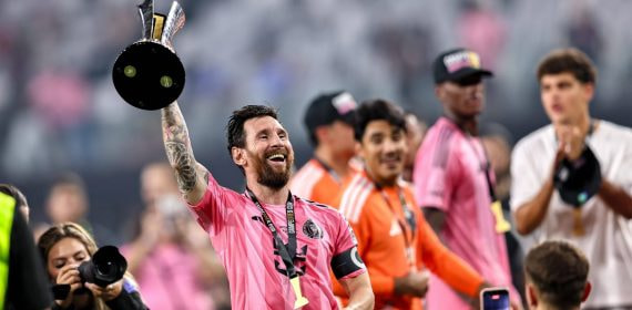Un futbolista con camiseta rosa, brazalete de capitán y medalla levanta un trofeo negro y plateado mientras sonríe