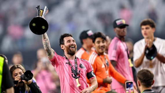 Un futbolista con camiseta rosa, brazalete de capitán y medalla levanta un trofeo negro y plateado mientras sonríe