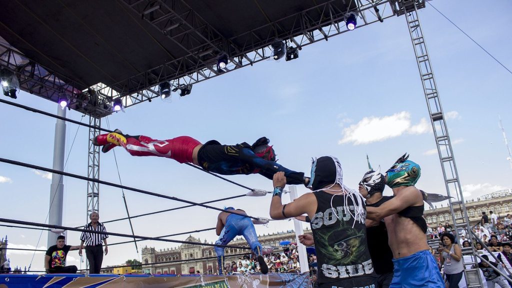 Un luchador enmascarado se lanza en vuelo sobre las cuerdas del ring mientras tres rivales lo esperan abajo, en una función al aire libre