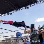 Un luchador enmascarado se lanza en vuelo sobre las cuerdas del ring mientras tres rivales lo esperan abajo, en una función al aire libre