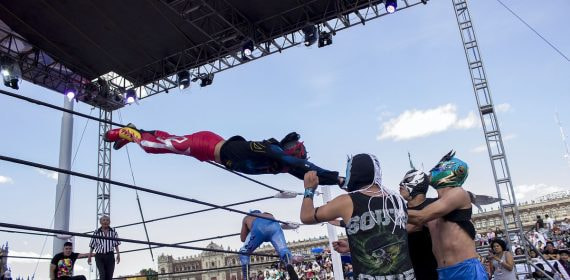 Un luchador enmascarado se lanza en vuelo sobre las cuerdas del ring mientras tres rivales lo esperan abajo, en una función al aire libre
