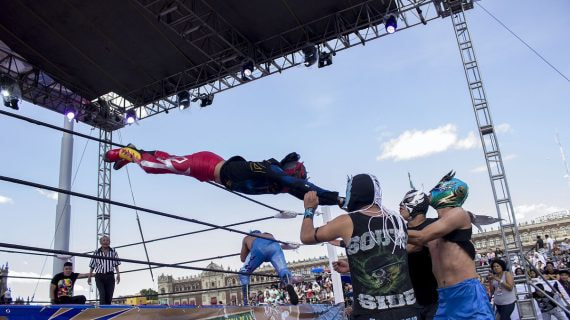 Un luchador enmascarado se lanza en vuelo sobre las cuerdas del ring mientras tres rivales lo esperan abajo, en una función al aire libre