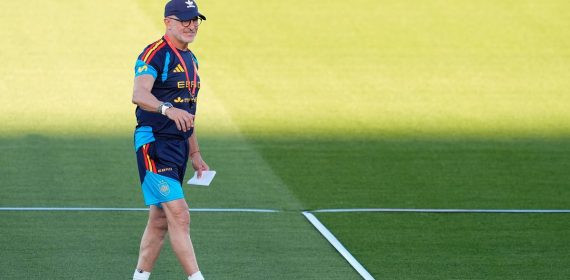 Luis de la Fuente, con gorra y ropa de entrenamiento de la selección, camina sonriente sobre el césped con un papel en la mano
