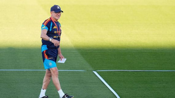 Luis de la Fuente, con gorra y ropa de entrenamiento de la selección, camina sonriente sobre el césped con un papel en la mano