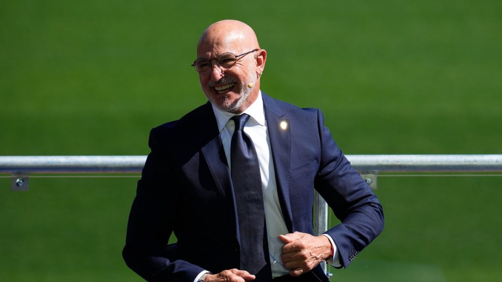 Luis de la Fuente, con traje azul marino y corbata, sonríe sentado junto a una barandilla sobre el césped de la Ciudad del Fútbol