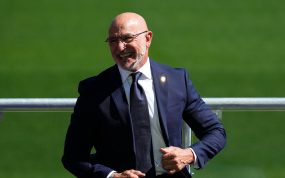 Luis de la Fuente, con traje azul marino y corbata, sonríe sentado junto a una barandilla sobre el césped de la Ciudad del Fútbol
