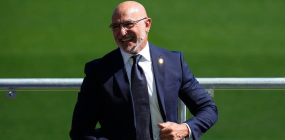 Luis de la Fuente, con traje azul marino y corbata, sonríe sentado junto a una barandilla sobre el césped de la Ciudad del Fútbol