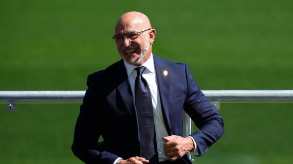 Luis de la Fuente, con traje azul marino y corbata, sonríe sentado junto a una barandilla sobre el césped de la Ciudad del Fútbol