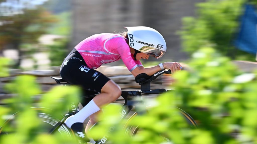 Una ciclista con casco aerodinámico blanco y maillot rosa pedalea acoplada en su bicicleta de contrarreloj entre vegetación