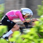 Una ciclista con casco aerodinámico blanco y maillot rosa pedalea acoplada en su bicicleta de contrarreloj entre vegetación