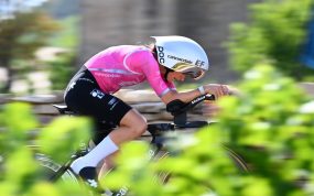Una ciclista con casco aerodinámico blanco y maillot rosa pedalea acoplada en su bicicleta de contrarreloj entre vegetación