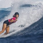 Una surfista con casco negro y licra rosa baja en cuclillas por la pared de una ola azul con la espuma detrás