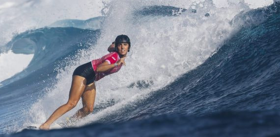 Una surfista con casco negro y licra rosa baja en cuclillas por la pared de una ola azul con la espuma detrás
