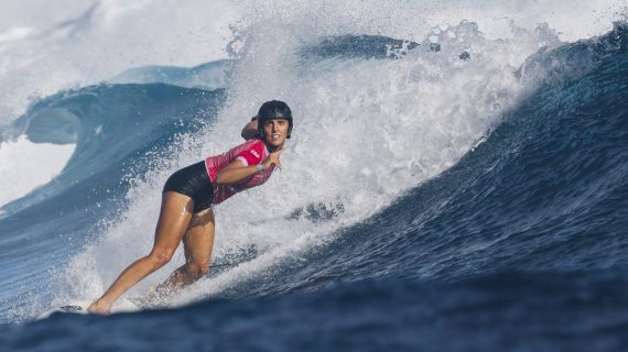 Una surfista con casco negro y licra rosa baja en cuclillas por la pared de una ola azul con la espuma detrás