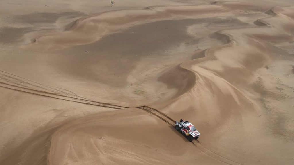 Un coche de rally blanco y rojo avanza entre grandes dunas de arena vistas desde el aire, dejando rodadas detrás