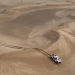 Un coche de rally blanco y rojo avanza entre grandes dunas de arena vistas desde el aire, dejando rodadas detrás