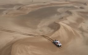 Un coche de rally blanco y rojo avanza entre grandes dunas de arena vistas desde el aire, dejando rodadas detrás