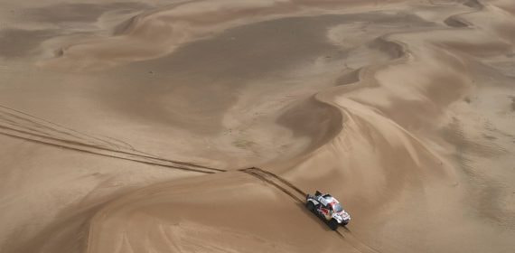 Un coche de rally blanco y rojo avanza entre grandes dunas de arena vistas desde el aire, dejando rodadas detrás