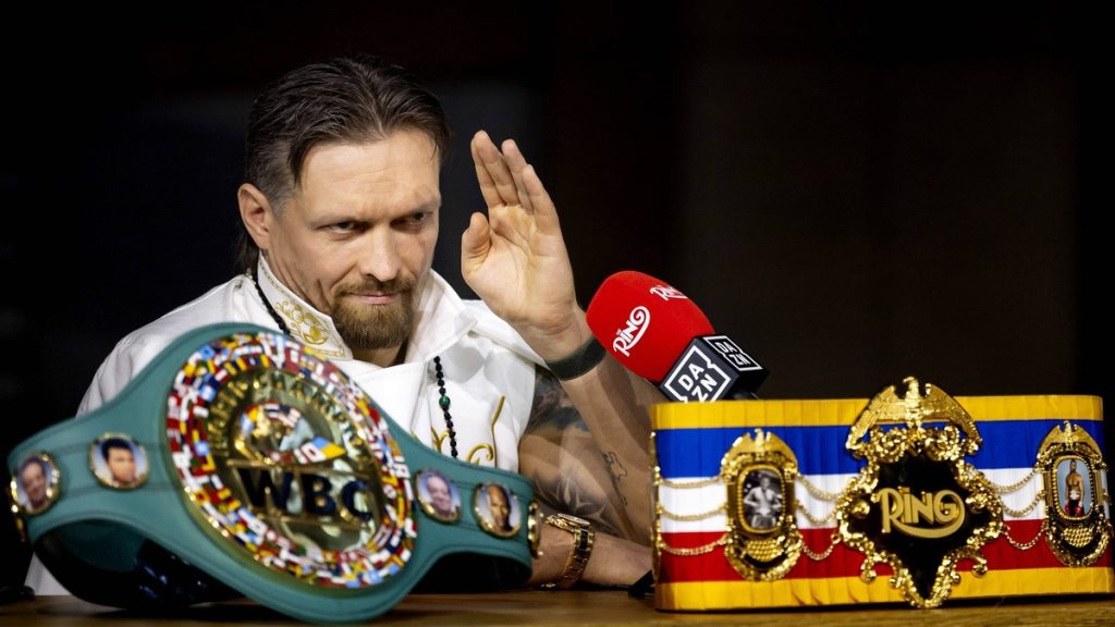 Oleksandr Usyk levanta la mano ante un micrófono, con el cinturón verde del CMB y el de The Ring sobre la mesa