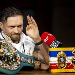 Oleksandr Usyk levanta la mano ante un micrófono, con el cinturón verde del CMB y el de The Ring sobre la mesa
