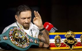 Oleksandr Usyk levanta la mano ante un micrófono, con el cinturón verde del CMB y el de The Ring sobre la mesa