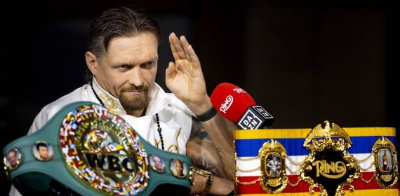 Oleksandr Usyk levanta la mano ante un micrófono, con el cinturón verde del CMB y el de The Ring sobre la mesa