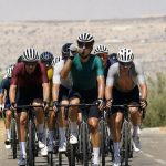 Un pelotón de ciclistas rueda de frente entre olivares resecos bajo un sol de mediodía mientras uno de ellos bebe de su bidón