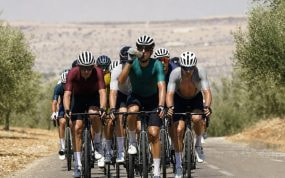 Un pelotón de ciclistas rueda de frente entre olivares resecos bajo un sol de mediodía mientras uno de ellos bebe de su bidón