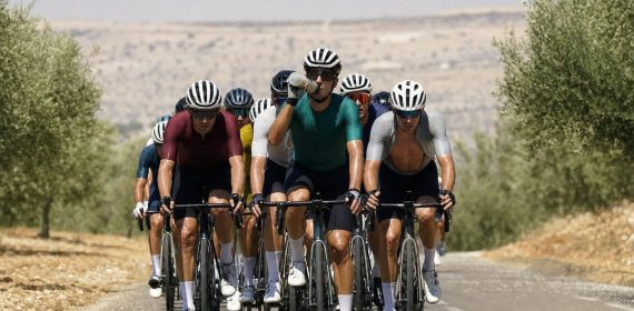 Un pelotón de ciclistas rueda de frente entre olivares resecos bajo un sol de mediodía mientras uno de ellos bebe de su bidón