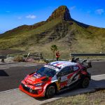 Un coche de rally rojo y blanco toma una curva de asfalto con una montaña de cima rocosa y el cielo azul al fondo