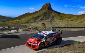 Un coche de rally rojo y blanco toma una curva de asfalto con una montaña de cima rocosa y el cielo azul al fondo