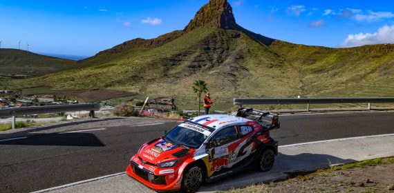Un coche de rally rojo y blanco toma una curva de asfalto con una montaña de cima rocosa y el cielo azul al fondo