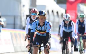 Un ciclista con el maillot celeste de Bélgica y gafas doradas rueda en cabeza de un grupo junto a las vallas del circuito