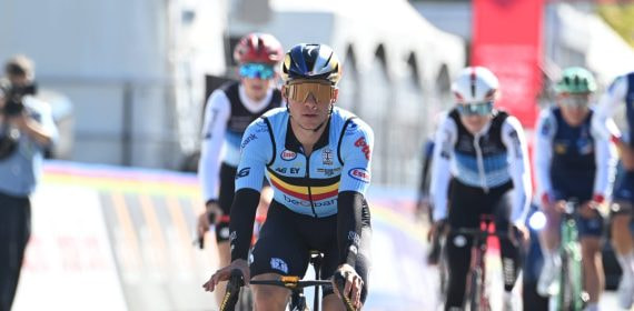 Un ciclista con el maillot celeste de Bélgica y gafas doradas rueda en cabeza de un grupo junto a las vallas del circuito