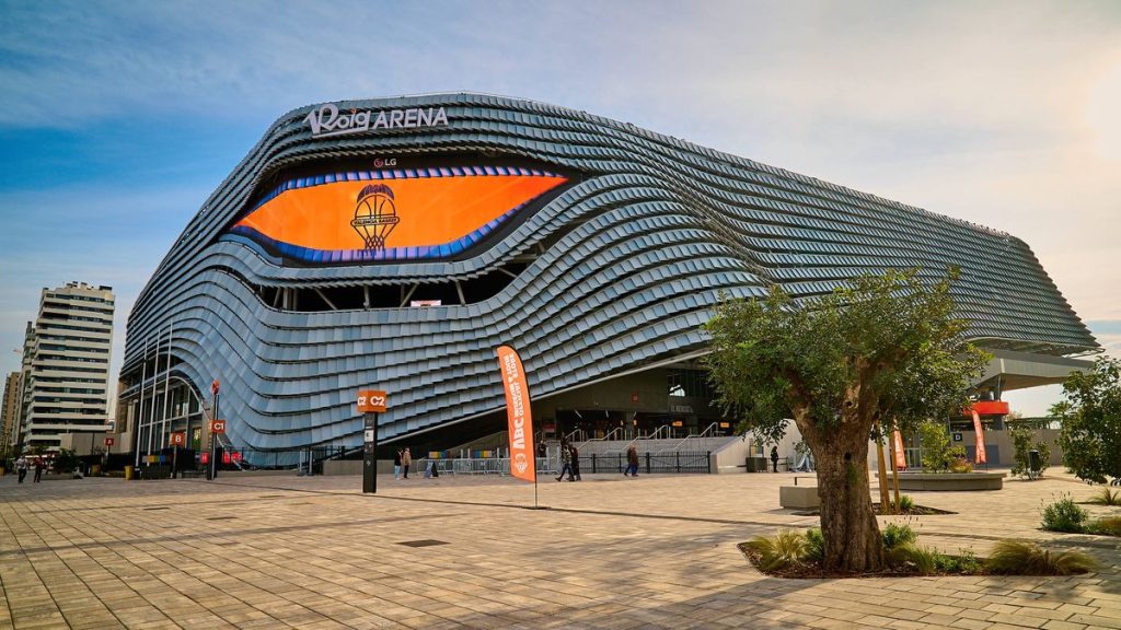 Fachada del Roig Arena, con placas plateadas onduladas y una pantalla naranja con el escudo del Valencia Basket, vista desde la explanada