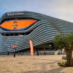 Fachada del Roig Arena, con placas plateadas onduladas y una pantalla naranja con el escudo del Valencia Basket, vista desde la explanada