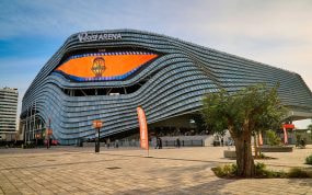 Fachada del Roig Arena, con placas plateadas onduladas y una pantalla naranja con el escudo del Valencia Basket, vista desde la explanada