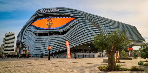 Fachada del Roig Arena, con placas plateadas onduladas y una pantalla naranja con el escudo del Valencia Basket, vista desde la explanada