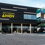 Fachada negra del pabellón Rotterdam Ahoy con su rótulo amarillo y una columna publicitaria delante de la entrada acristalada