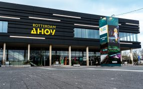 Fachada negra del pabellón Rotterdam Ahoy con su rótulo amarillo y una columna publicitaria delante de la entrada acristalada