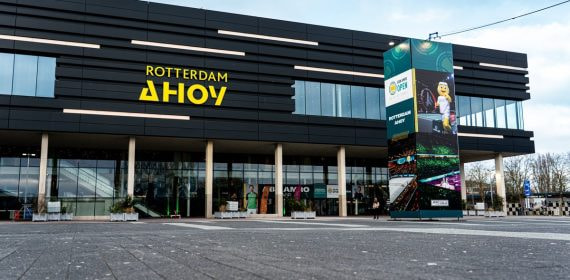 Fachada negra del pabellón Rotterdam Ahoy con su rótulo amarillo y una columna publicitaria delante de la entrada acristalada
