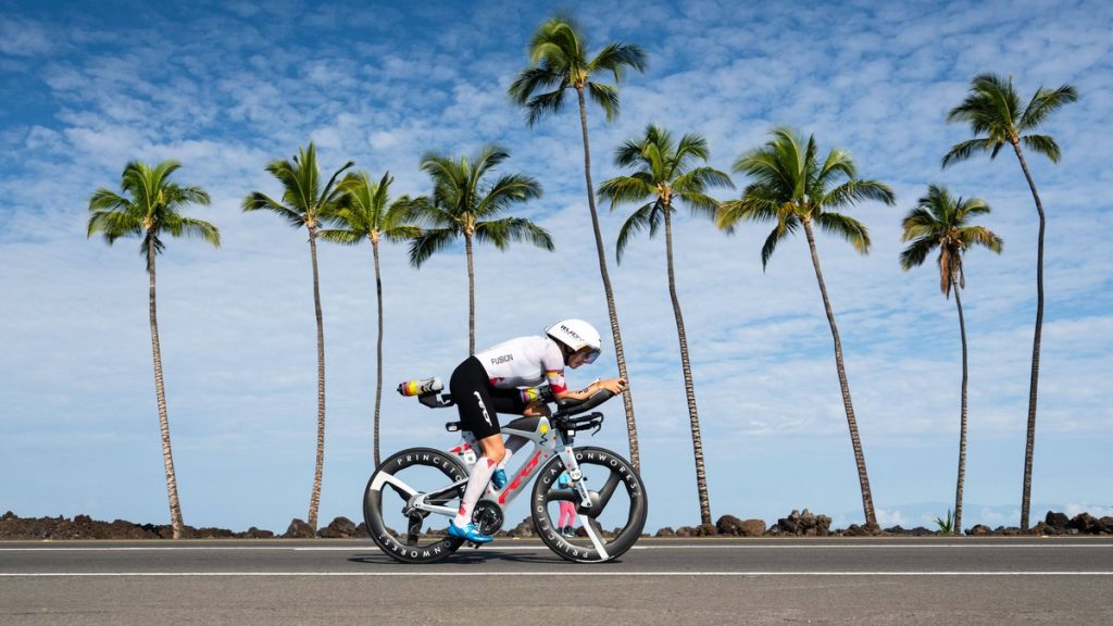 Una triatleta con casco blanco pedalea acoplada en su bicicleta de contrarreloj por una carretera flanqueada de palmeras