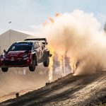 Un Toyota GR Yaris Rally1 rojo y blanco salta sobre un tramo de tierra entre una nube de polvo y llamaradas