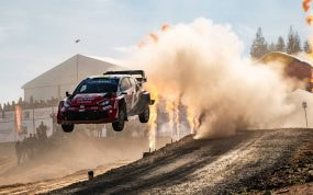 Un Toyota GR Yaris Rally1 rojo y blanco salta sobre un tramo de tierra entre una nube de polvo y llamaradas