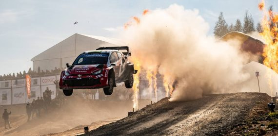Un Toyota GR Yaris Rally1 rojo y blanco salta sobre un tramo de tierra entre una nube de polvo y llamaradas