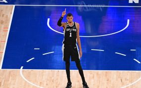 Victor Wembanyama, con la camiseta negra de los Spurs, levanta el dedo índice de pie en la zona de la pista